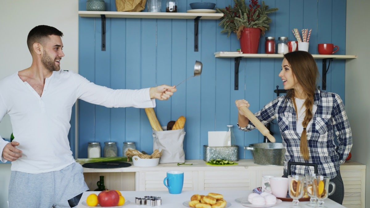 Happy couple having fun in the kitchen fencing with ladle and spoon while cooking breakfast at home in the morning