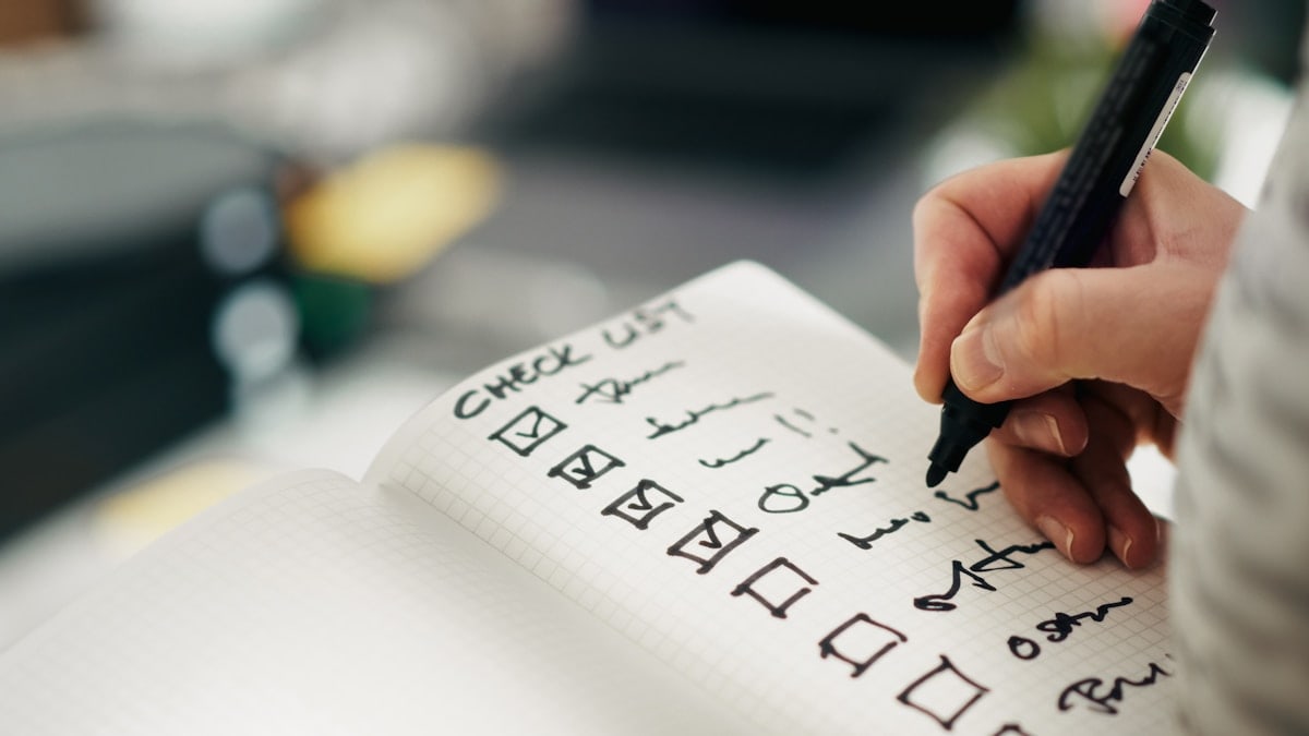 Checklist notebook task progress accuracy. Close-up hand holding pen while marking completed tasks grid notebook. Symbol of productivity, precision, quality control, and workflow management in business or personal planning